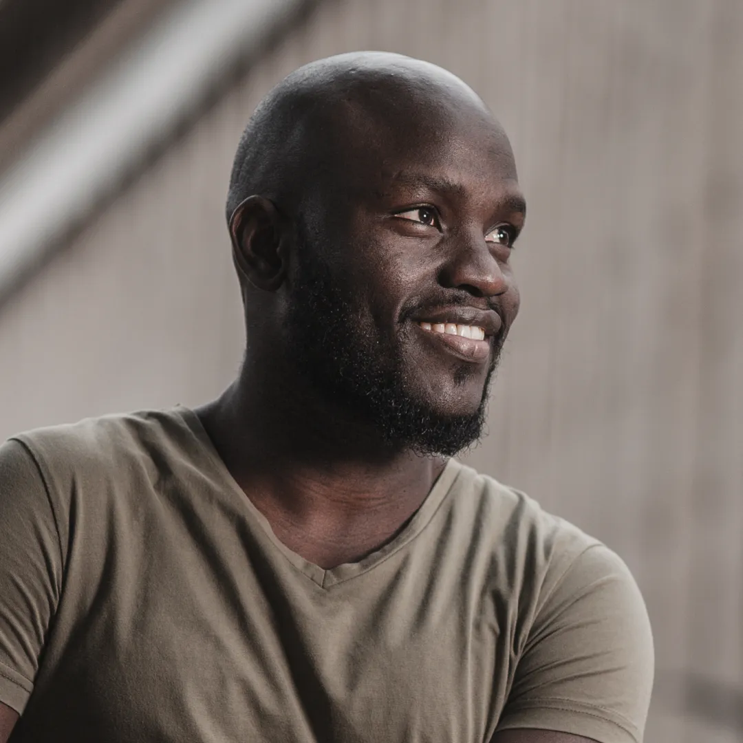 A man in an olive v-neck t-shirt smiles warmly while looking off to the side against a neutral background.